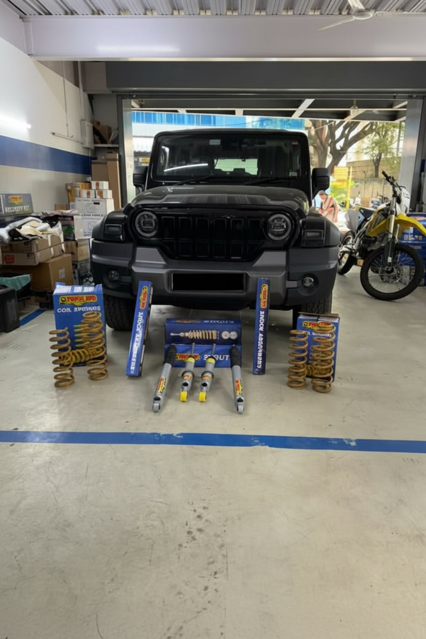 A set of blue and yellow coilover suspension components, including springs, shock absorbers, and knuckle assemblies, displayed on the ground in front of a black SUV inside a garage workshop.
