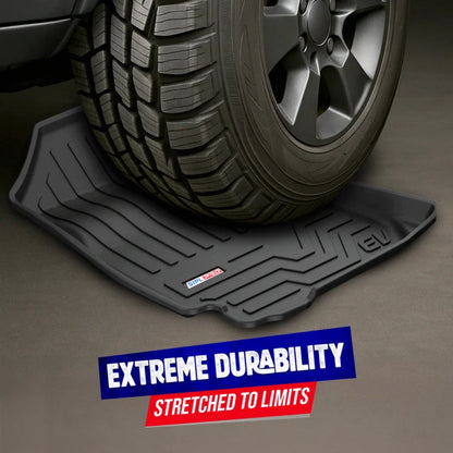 A black rubber vehicle floor mat shown under a tire on a gray floor, with a blue and red text banner at the bottom reading 'EXTREME DURABILITY' and 'STRETCHED TO LIMITS'.