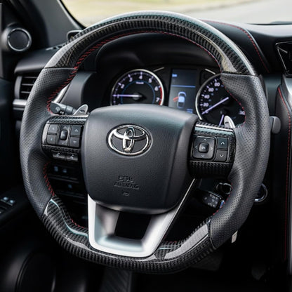 A close-up of a Toyota steering wheel with a black carbon-fiber-look wrap and red contrast stitching, shown inside a vehicle cabin.