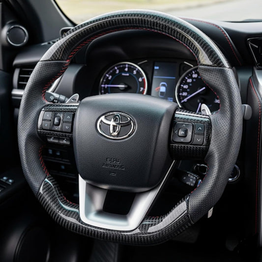 A close-up of a Toyota steering wheel with a black carbon-fiber-look wrap and red contrast stitching, shown inside a vehicle cabin.