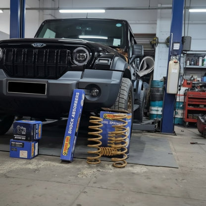 A set of car suspension parts, including a gold coil spring and blue shock absorbers in their retail boxes, displayed on the floor next to a black vehicle in an auto repair shop.